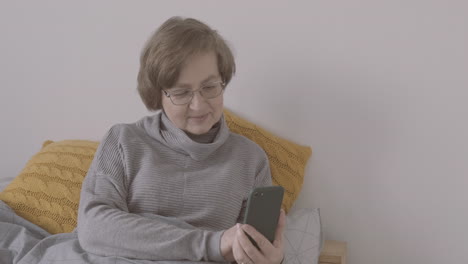 ältere-Frau-Mit-Brille,-Die-Mit-Smartphone-Auf-Dem-Bett-Sitzt