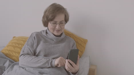 Senior-Woman-With-Eyeglasses-Sitting-On-Bed-Making-A-Video-Call-On-A-Smartphone