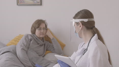Senior-Woman-Sitting-On-Bed-While-Talking-To-Female-Doctor-In-Medical-Mask-And-Protective-Screen-Using-A-Table