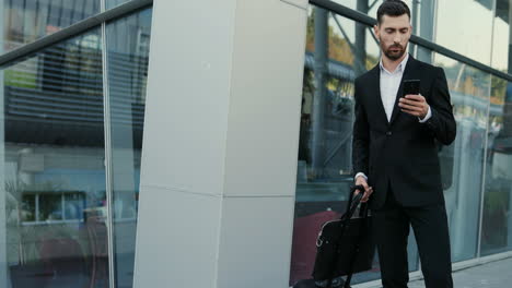 Good-Looking-Businessman-In-Suit-Standing-Outdoor-With-His-Suitcase-And-Tapping-Or-Scrolling-On-The-Smartphone