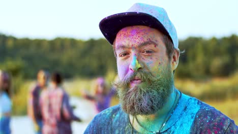 Close-Up-Of-The-Young-Handsome-Man-With-A-Beard-And-In-A-Hat-Turning-Face-Tothe-Camera-And-Smiling-While-Being-In-Colorful-Spots-Of-Paints-As-Celebrating-Holi-Festival-With-Friends