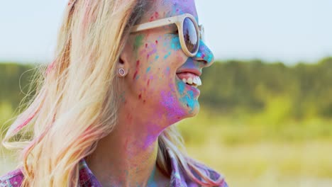 Close-Up-Of-The-Young-Blond-Beautiful-Woman-In-Sunglasses-Smiling-And-Looking-At-The-Side-While-Her-Face-Being-In-Colorful-Paints-As-She-Celebrating-Holi,-Holiday