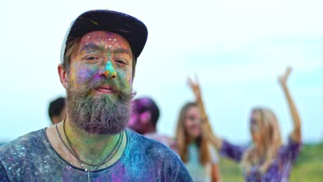 Portrait-Of-The-Stylish-Hipster-Guy-In-Hat-And-With-Beard-And-Moustaches-Looking-At-The-Side,-Then-Smiling-To-The-Camera-While-Being-All-In-Colorful-Paints-At-The-Holi-Fest