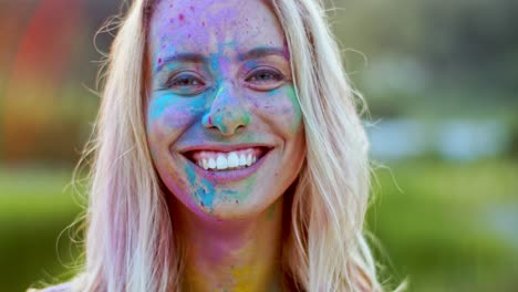 Portrait-Shot-Of-The-Pretty-Charming-Blond-Woman-With-Colorful-Paints-On-Her-Face-Smiling-Cheerfully-To-The-Camera-Outdoor-At-The-Holi-Festival