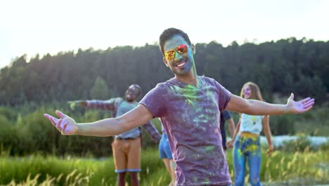Portrait-Of-The-Young-Handsome-Man-In-Sunglasses-And-All-In-Colorful-Paints-Spots-Smiling-To-The-Camera-And-Posing-Outdoors-While-Having-Festive-Holi-Day-With-Multiethnic-Friends