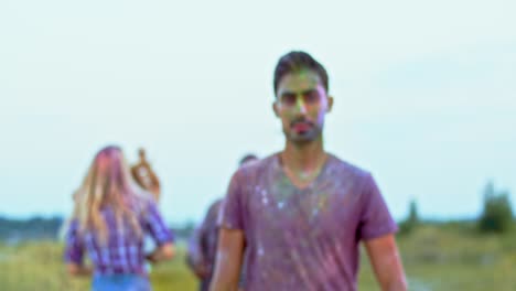 Blurred-Shot-Of-The-Young-People-Dancing-And-Having-Fun-Outdoors-At-The-Holi-Festival,-Handsome-Young-Man-Coming-Closer-To-Camera,-Smiling-And-Posing-To-The-Camera