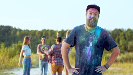 Portrait-Shot-Of-The-Young-Attractive-Sporty-Man-With-Beard-And-In-A-Hat-Looking-Back-At-His-Multiethnic-Friends-Who-Dancing-And-Having-Fun-With-Paints-During-Holi-Festival,-Then-Turning-Face-To-The-Camera-And-Smiling-To-The-Camera