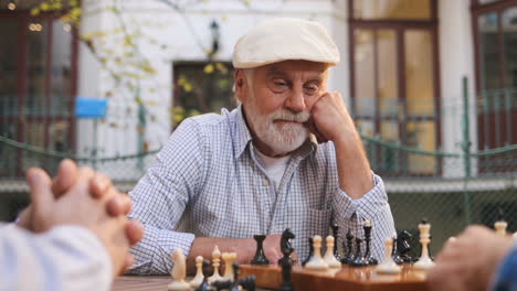 Porträt-Des-Alten-Mannes-Mit-Mütze,-Der-Mit-Freunden-An-Der-Frischen-Luft-Sitzt-Und-An-Die-Nächste-Runde-Im-Schachspiel-Denkt