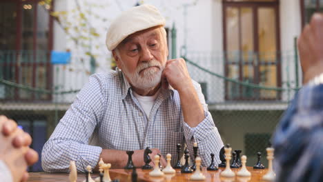 Portrait-Shot-Of-The-Grey-Haired-Good-Looking-Man-On-Retirement-Sitting-At-The-Table-On-A-Fresh-Air,-Leaning-On-His-Arm-And-Looking-At-The-Friend's-Hand-Moving-A-Chessman-On-A-Board