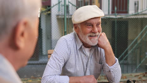 Close-Up-Of-The-Wise-Old-Man-In-A-Cap-Sitting-At-The-Chess-Game-With-Friends-And-Talking-While-Considering-Next-Turn