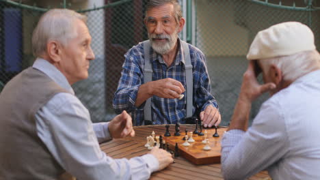 Portrait-Shot-Of-The-Three-Old-Grandfathers-Playing-Chess-Game-In-The-Yard-Outdoors,-One-Man-Moving-His-Figure-And-Others-Feeling-Like-He-Winning