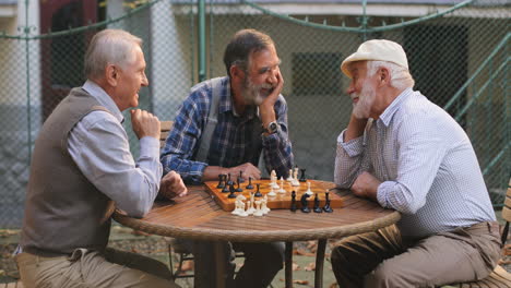 Porträtaufnahme-Der-Drei-älteren,-Fröhlichen,-Freundlichen-Männer-Im-Ruhestand,-Die-Mit-Einem-Schachspiel-Im-Hof-Um-Den-Tisch-Sitzen-Und-Lächeln