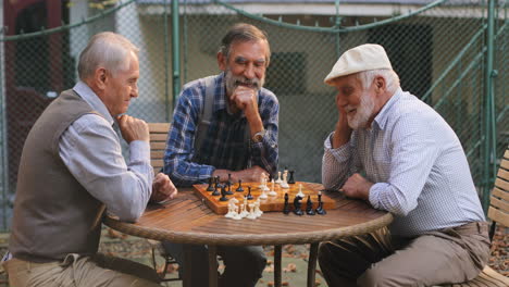 Porträt-Der-Drei-Alten-Glücklichen-Männlichen-Freunde-Im-Ruhestand,-Die-Bei-Einem-Schachspiel-Im-Hof-Am-Tisch-Sitzen-Und-Beim-Nachdenken-Lächeln