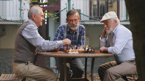 Porträtaufnahme-Der-Drei-Senioren-Im-Ruhestand,-Die-Im-Hof-Um-Den-Tisch-Herum-Sitzen-Und-Eine-Schachpartie-Spielen,-Während-Sie-Figuren-Bewegen