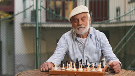 Portrait-Shot-Of-The-Old-Grey-Haired-Clever-Man-In-A-Cap-Sitting-At-The-Table-In-Front-Of-The-Chess-Board-And-Moving-A-Figure,-Then-Smiling-To-The-Camera
