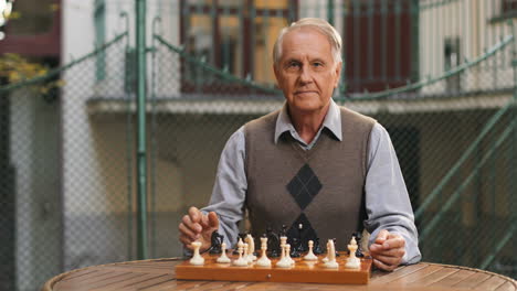 Close-Up-Of-The-Old-Retired-Wise-Man-Sitting-In-The-Yard-At-The-Table-With-The-Chess-Game-On-And-Looking-At-The-Camera