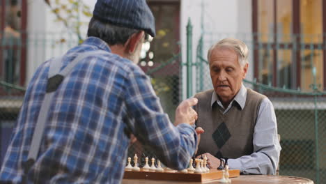 Back-View-On-The-Senior-Grandfather-In-A-Cap-Sitting-In-His-Yard-With-Old-Neighbor-And-Talking-While-Playing-Chess