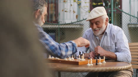 View-Over-The-Tree-On-The-Two-Male-Old-Cheerful-Friends-Sitting-On-A-Fresh-Air-In-The-Yard-And-Playing-Chess