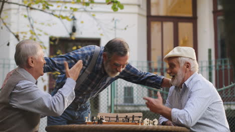 Two-Old-Men-Playing-Chess-Game-In-The-Yard-And-Their-Friend-Coming-To-Them,-Greeting-And-Shaking-Hands