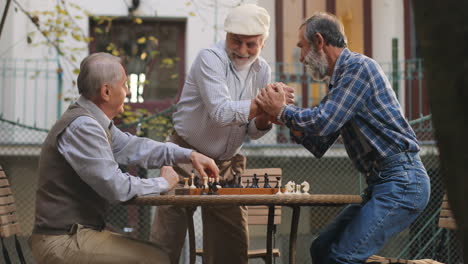 Fröhlicher-älterer-Mann-Mit-Mütze,-Der-In-Den-Hof-Kommt-Und-Sich-Seinen-Beiden-Nachbarn-Beim-Schachspiel-Anschließt