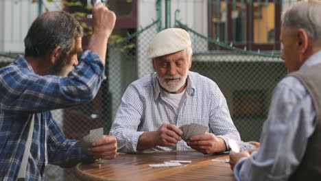 Three-Senior-Men-Best-Friends-On-Retirement-Playing-Cards-In-The-Yard-At-The-Table