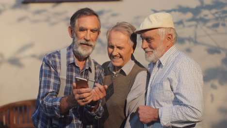 Ältere-Männliche-Freunde-Im-Ruhestand-Stehen-Im-Freien-Und-Sehen-Sich-Online-Etwas-Auf-Dem-Smartphone-Bildschirm-An