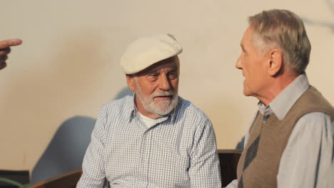Close-Up-Of-The-Two-Old-Retired-Men-Neighbors-Talking-While-Sitting-And-Resting-On-The-Bench-On-A-Sun-Their-Friend-Coming-To-Them-And-Shaking-Hands
