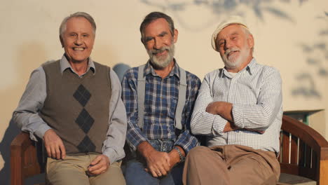 Porträt-Der-Drei-Alten-Männer,-Die-Auf-Einer-Bank-Sitzen-Und-Mit-Einem-Lächeln-An-Der-Wand-In-Die-Kamera-Posieren