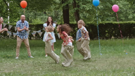Children-Playing-To-Sack-Race-At-A-Birthday-Party-In-The-Park-As-Their-Parents-Cheer-Them-On