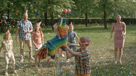 Niños-Con-Sus-Padres-En-Una-Fiesta-De-Cumpleaños-En-El-Parque,-En-Primer-Plano-Un-Niño-Rompe-Una-Piñata