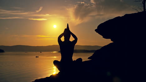 Backlit-View-Of-A-Woman-Doing-Yoga-Poses-Sitting-On-A-Stone-In-Front-Of-The-Sea-At-Sunset