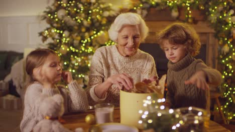 Großmutter-Mit-Zwei-Enkelkindern,-Die-Am-Tisch-Sitzen-Und-Geschenke-In-Einem-Raum-Mit-Weihnachtsdekoration-Verpacken