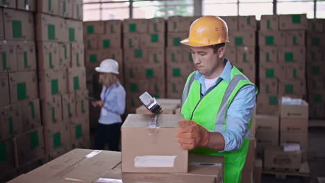 Warehouse-Worker-Wearing-A-Helmet-And-Reflective-Vest-Seals-A-Box-With-Duct-Tape,-In-The-Blurred-Background-A-Woman-Is-Placing-Cardboard-Boxes-On-The-Shelf