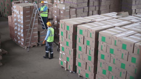 Two-Warehouse-Men-Wearing-Helmets-And-Reflective-Vests,-One-Of-Them-Places-Cardboard-Boxes-And-The-Other-Carries-A-Wheelbarrow