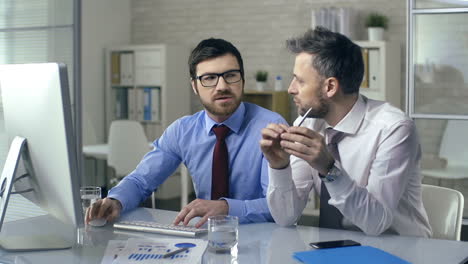 Zwei-Geschäftsmänner-In-Hemden-Und-Krawatten-Sprechen-über-Ein-Projekt-Und-Schauen-Auf-Den-Computerbildschirm,-Der-An-Einem-Tisch-Im-Büro-Sitzt