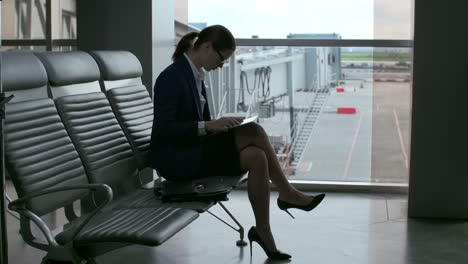 Business-Woman-In-Glasses,-Suit-And-Heels-Using-A-Tablet-While-Sitting-In-The-Airport-Looking-Out-The-Window-While-Waiting-For-A-Flight