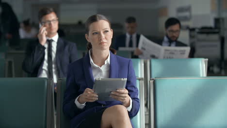 Business-Woman-In-Suit-Using-A-Tablet-While-Sitting-In-The-Airport-Waiting-Area,-Behind-Are-Several-Men-In-Suits-Sitting