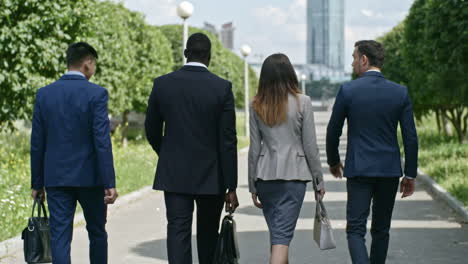 Businessmen-And-Businesswoman-Wearing-Elegant-Suits-Talking-And-Holding-Briefcases-While-They-Walking-In-The-Park