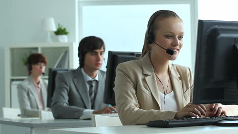 Telemarketer-Office,-In-The-Foreground-A-Blonde-Woman-Talking-With-A-Headset-And-Typing-On-The-Computer-Keyboard