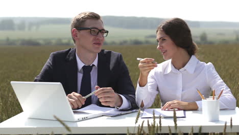 Businesswoman-And-Businessman-Sitting-At-A-Table-In-Front-Of-The-Laptop-While-Talking-And-Laughing-In-A-Wheat-Field