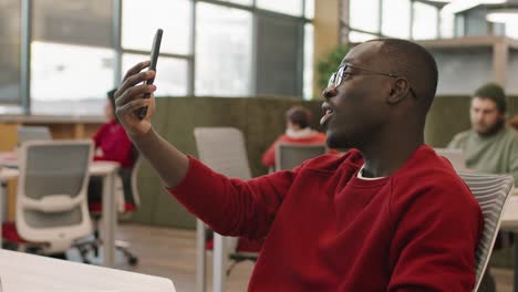 Arbeiter-Mit-Brille-Und-Rotem-Pullover-Sitzen-Im-Büro,-Während-Sie-Einen-Videoanruf-Mit-Dem-Smartphone-Tätigen