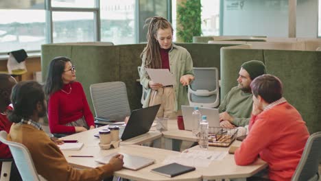 Meeting-Of-Coworkers-Sitting-At-A-Table-In-The-Office,-Woman-With-Dreadlocks-Stand-Talk-About-A-Project