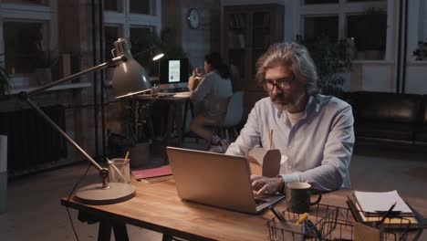Grauhaariger-Mann-Mit-Brille-Und-Blauem-Hemd-Sitzt-Vor-Dem-Computer,-Während-Er-Isst-Und-Arbeitet,-Im-Hintergrund-Arbeitet-Sein-Partner-An-Einem-Schreibtisch-Sitzend