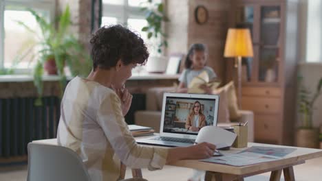 Woman-With-Short-Curly-Hair-Sitting-At-A-Desk-In-Front-Of-The-Computer-While-Presenting-A-Project,-In-The-Background-His-Daughter-Is-Reading-A-Book