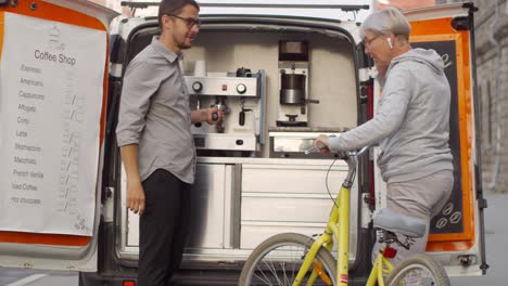 Blonde-Woman-Riding-A-Bicycle,-Stops-In-Front-Of-A-Cafe-Truck-And-Asks-The-Waiter-For-A-Coffee