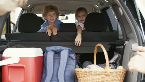 Family-Goes-On-Vacation,-Parents-Put-Luggage-In-The-Trunk-And-Little-Children-Wait-Sitting-Inside-The-Car