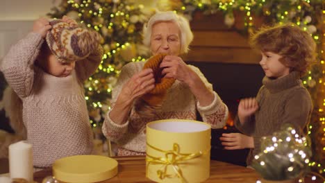 Grandmother-And-Grandchildren-Open-Gift-At-Christmas,-Grandmother-Puts-Woolly-Hats-On-Her-Grandchildren
