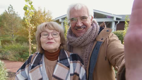 Cheerful-Senior-Man-And-Woman-In-Stylish-Warm-Casualwear-Looking-At-You-With-Smiles-While-Greeting-To-Camera-Against-Garden-And-House