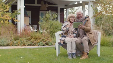 Older-Couple-Sitting-On-A-Bench-In-Their-Home-Garden-While-Talking-On-A-Video-Call-Using-A-Tablet