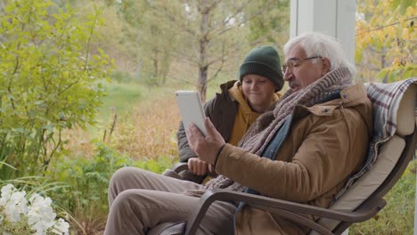 Senior-Grandfather-Sitting-With-His-Grandson-Sitting-On-A-Bench-In-Home-Garden-While-Talking-On-A-Video-Call-Using-A-Tablet